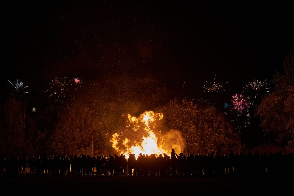 Large crowd of people circle around huge bonfire with fireworks in background, lit at night