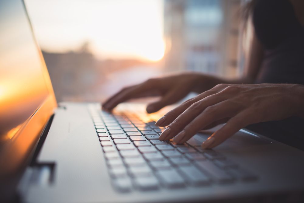 Up close view of person's hands typing at a laptop computer, reflecting the sunset