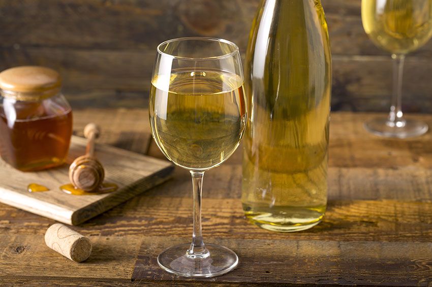 A honey pot, two glasses of mead, and a bottle of mead on a wooden table.