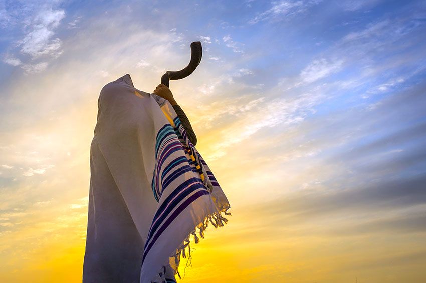 Jewish man in a traditional tallit prayer shawl blowing the ram's horn against dramatic sunset sky.